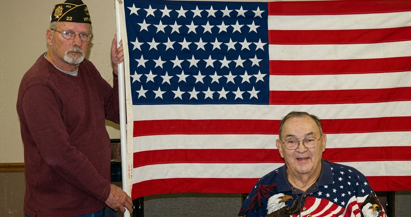 Two older men pose indoors with a large American flag; one standing in a maroon sweater and cap, the other seated in a patriotic shirt with stars and stripes.